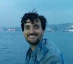 Smiling white male with scraggly moustache and beard and windswept hair with the Turkish strait behind.